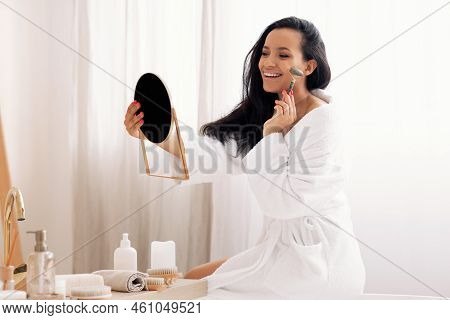 Female Massaging Face With Roller Looking At Mirror In Bathroom