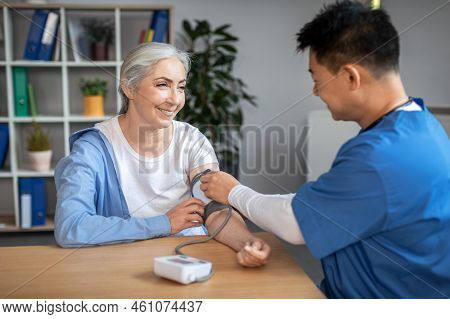 Glad Young Asian Man Doctor In Uniform Measures Blood Pressure With Tonometer To Elderly European Wo