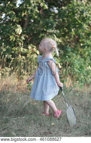 Blonde Little Girl With Two Tails And Summerdress Plays Badminton. Child Holds Racket And Shuttlecoc