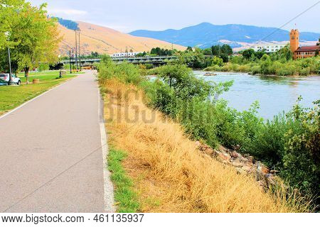Pedestrian Riverwalk Besides The Clark Fork River With The Historical Train Depot And Mountains Beyo