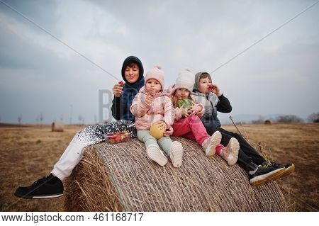 Four Kids With Fruits In Hands Sitting On Haycock At Field.