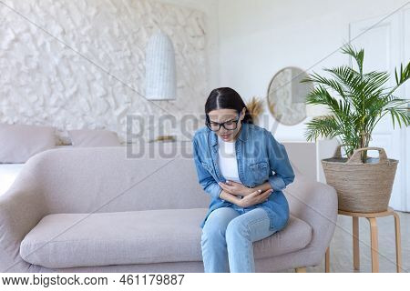 Stomach Ache. Young Beautiful Woman In Glasses And Jeans Sitting On The Sofa At Home And Holding Her