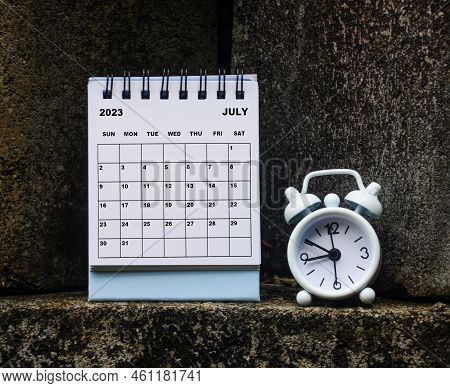 July 2023 White Desk Calendar With Alarm Clock Glass On Rock Background. Calendar Concept.