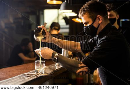 A Bartender In A Black Protective Mask Pours A Cocktail From A Viker Into A Chilled Glass Through A 