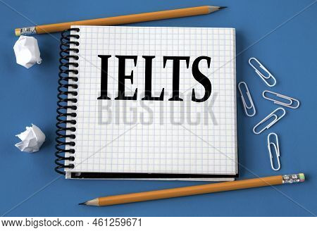 Ielts - Word In A White Notebook On A Blue Background With Pencils, Paper Clips. Info Concept