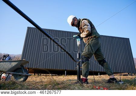 Male Worker Building Pile Foundation For Wooden Frame House. Man Builder In White Safety Helmet Dril