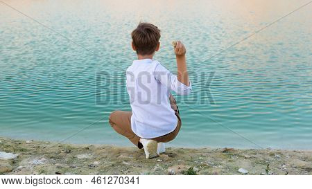 A Boy 8-9 Years Old Throws Stones Into The Water From The Shore, Fun In Nature.