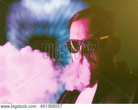Pensive Bearded Man Dressed In A Leather Jacket, Sunglasses Vaping. A Cloud Of Vapor.