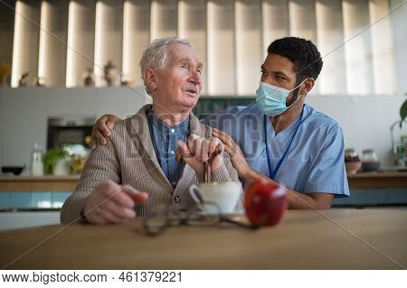 Caregiver With Medical Mask Bringing Healthy Snack To Senior Man In Nursing Home Care Center.