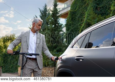 Businessman In Suit On Way To Work Standing At Eletric Scooter And Charging His Electric Car. Concep