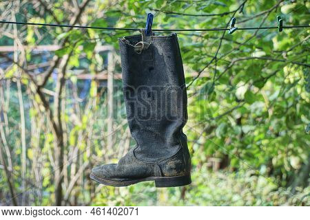 One Old Dirty Black Tarpaulin Boot Hangs On A Wire On The Street Against The Background Of Green Veg