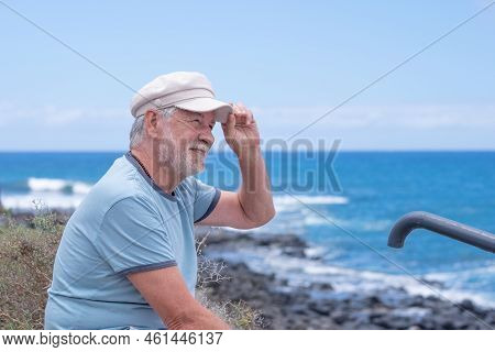 Smiling Caucasian Bearded Senior Man With Hat Sitting At The Beach Looking At The Horizon While Enjo
