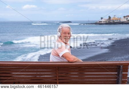 Cheerful Senior Bearded Man Sitting On A Bench In Front To The Sea Relaxing Listening Music By Headp