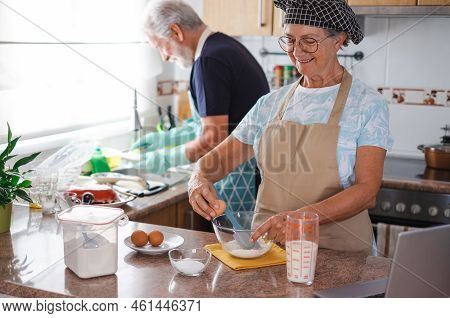 Smiling Senior Woman Preparing Her Homemade Plumcake While Her Husband Washes The Dishes. Concept Of