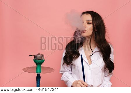 Young, Attractive Woman Smokes A Hookah, Sheesha On A Pink Background. The Pleasure Of Smoking.