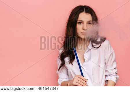 Young Woman Smokes A Hookah, Sheesha On A Pink Background. The Pleasure Of Smoking.