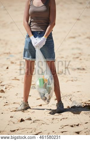 Cropped Image Of Young Woman Standing On Sandy Beach With Plastic Bag Of Garbage She Found
