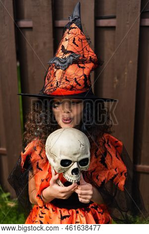 Curly Girl In Witch Hat Pouting Lips Near Spooky Skull Outdoors.