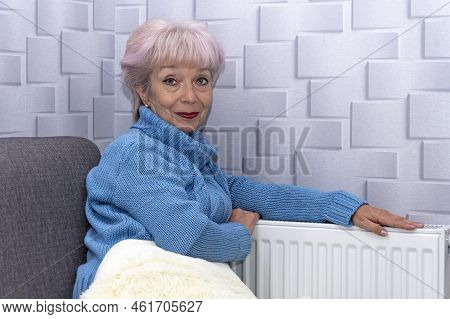 An Elderly Woman In A Blue Sweater Warming Her Hands On A Heating Radiator. Concept: Low Room Temper