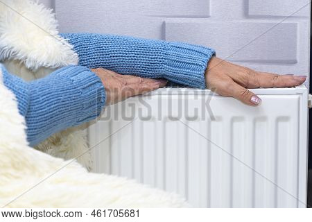 An Elderly Woman In A Blue Sweater Warming Her Hands On A Heating Radiator. Concept: Low Room Temper