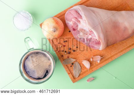 Top View Of Raw Pork Knuckle, Garlic, Onion, Dried Bay Leaves, Sea Salt And Peppercorns On Wooden Cu