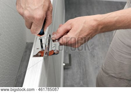 Man Installs The Door Hinge On The Door With A Screwdriver. Tightening The Screws. Renovation In The
