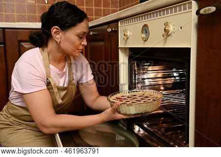 Middle-aged Beautiful Latin American Woman Wearing Beige Chef Apron, Cooking A Cherry Pie In The Ove
