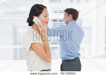 Side view of a smiling businesswoman using cellphone with man in background at office