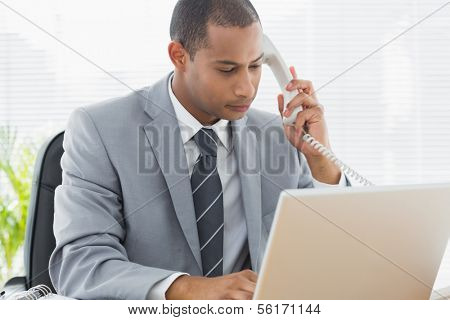 Concentrated young businessman using laptop and phone at office desk