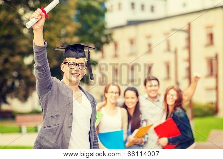 education, campus and teenage concept - smiling teenage boy in corner-cap and eyeglasses with diploma and classmates on the back