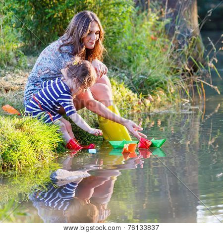 Adorable Little Girl And Her Mom Playing With Paper Boats In A River.