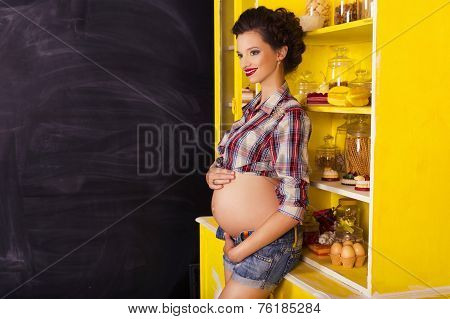 Beautiful Brunette Woman On A 7Th Month Pregnancy In Plaid Shirt And Jeans Shorts On A Yellow Kitche