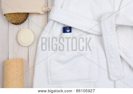 Closeup of a bathrobe with lotion in the pocket. A scrub brush and soap and a luffa are next to the robe on a rustic wooden surface.