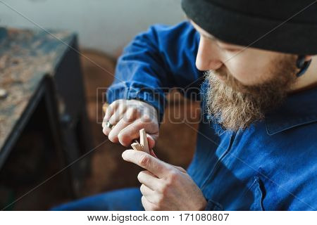 Man with a beard in blue jeans suit carving a wooden spoon with a knife, close up, woodworking, copy space.