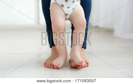 Bare legs of the child, dressed in a white diaper, standing on the bare soles of the feet of mother dressed in blue jeans, baby feet are between mothers legs, portrait on the background of the home environment.