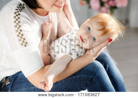 Cute young mother with thick black hair, dressed in a white T-shirt and blue jeans playing with her red-haired young son, dressed in a white shirt and white panties with gray stars, putting it on her lap, sitting on a bed in a light bedroom