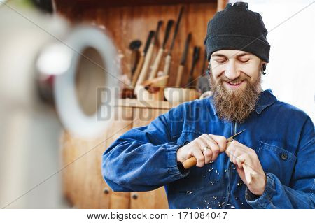 Worker with a beard wearing blue jeans suit and black hat carving wood and looking at wooden spoon, woodcarving instruments at background.
