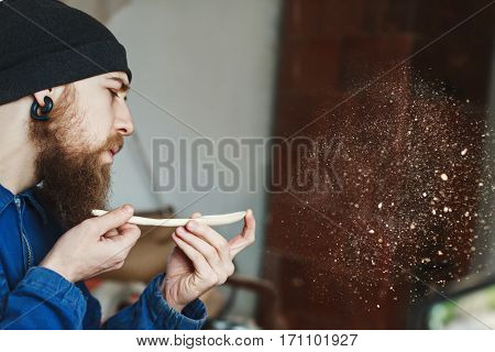 Worker with a beard wearing blue jeans suit and black hat holding wooden spoon and blowing dust, woodcarving, portrait, copy space, woodworking.