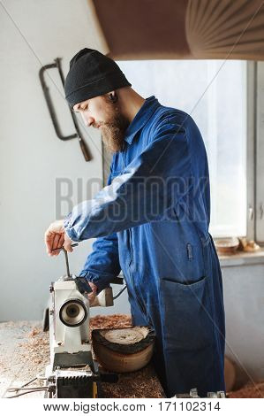 Man with a beard wearing blue jeans suit and black hat working near table, woodcarving instruments on table, big window at background, portrait, woodworking.