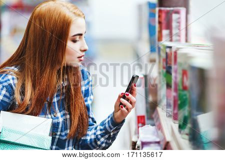 Beautiful young girl with long brown hair wearing blue plaid shirt shopping at store, holding boxes and mobile phone, copy space, making a photo.
