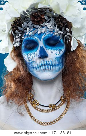 Santa Muerte make up. Young woman with short curly hair and creative visage. White flowers in hair, make up, portrait at blue background, wearing golden necklaces.