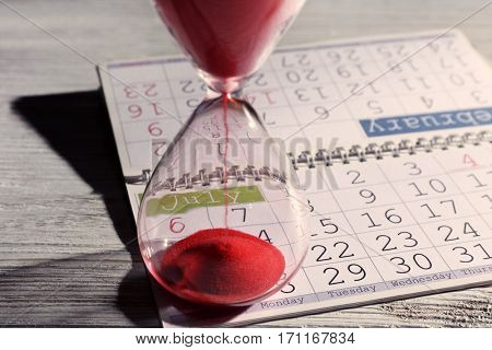 Time passing concept. Crystal hourglass with red sand and calendar on light wooden background