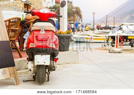 Red two-wheeled moped parked on Mithos beach promenade in sea bay of resort village Bali. Classic Greek stone architecture of resort tourists walk streets of city. Rethymno Crete Greece