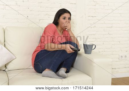 young beautiful latin woman sitting at home sofa couch in living room watching television scary horror movie or horrible news scared and excited covering her mouth holding remote control