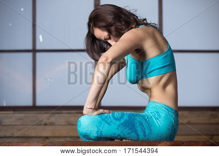 Young attractive woman practicing yoga, making Upward Abdominal Lock, Uddiyana Bandha, sitting in Half Lotus pose, working out, wearing sportswear, blue tank top, pants, full length, evening practice