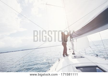 Senior man wearing jacket, scarf and hat, standing on bow of yacht, pulling rope adjusting mainsail during perfect autumn day under sails - sailing holidays concept