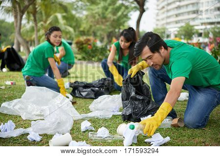 Portrait of middle-aged Asian man in casualwear crouching to plastic waste and picking it up in bin bag