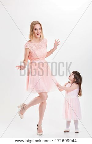 blonde smiling mother and cute little daughter in pink Princess dresses pulls her dress on a gray background. family