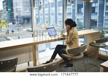 Young afroamerican executive making remote job being in business trip sitting at table in coworking space talking on the phone with entrepreneur using laptop computer with mock up screen with wifi