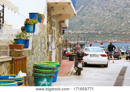 Bali Greece - April 30 2016: Two-wheeled moped and white car Audi A5 coupe parked on Mithos beach promenade in sea bay of resort village Bali. Classic Greek stone architecture of resort tourists walk streets of city. Rethymno Crete Greece